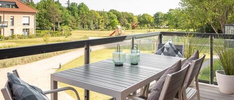 A balcony with a table and chairs, a potted plant, and a view of a playground and trees.