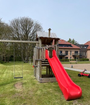A playground with a slide and swings, surrounded by grass and trees.