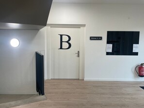 Ein Flur mit einer Tür, auf der 'B' steht, einem Schild mit der Bezeichnung Zimmer 0.1-0.4, einem Feuerlöscher und einem Heizkörper.