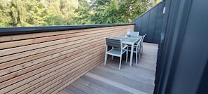 A wooden deck with a table and chairs, surrounded by a dark wall and a view of trees.