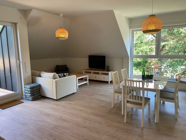 A modern living room with a sofa, TV, dining table, and chairs. There are two hanging lights and a large window with a view of greenery.