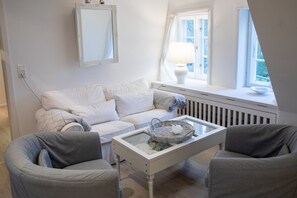 A room with a white sofa, two armchairs, a small table with a basket, a white radiator, and a window with a view. - Vacation apartment Silbermöwe- Herrenhof Wrixum - Vacation in the historic (Wyk auf Föhr)