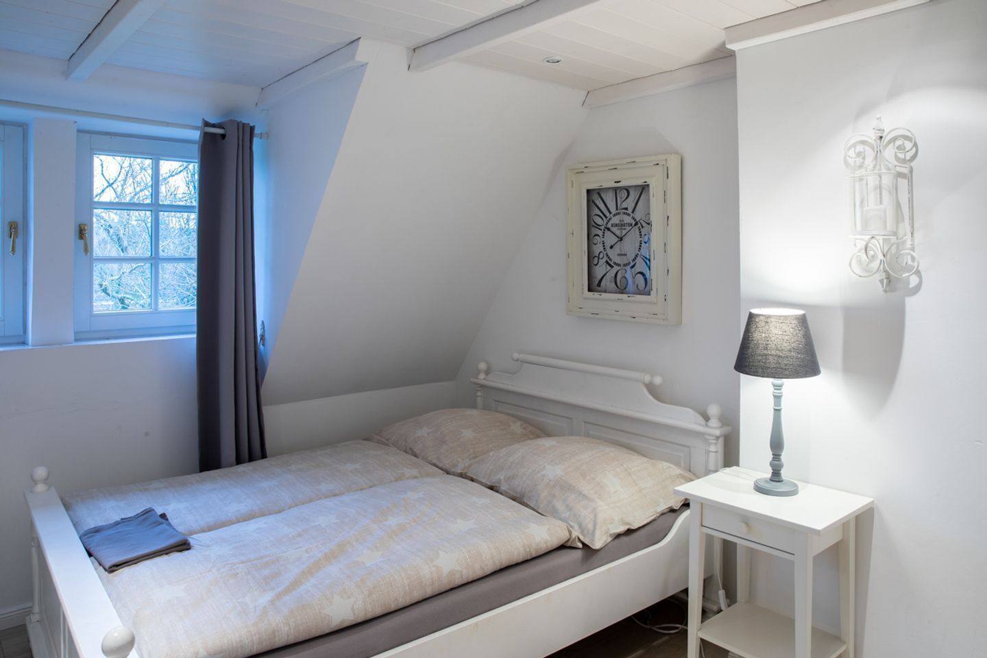 Une chambre à coucher avec un lit, une table de chevet avec une lampe, une fenêtre avec des rideaux et une horloge murale.
