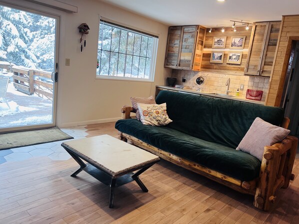A cozy living room with a wooden floor, a green sofa with cushions, a wooden coffee table, and a large window with a view of a snowy outdoor deck.