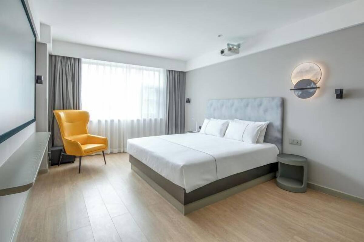 A modern hotel room with a bed, a chair, a bedside table, a lamp, and a window with curtains.
