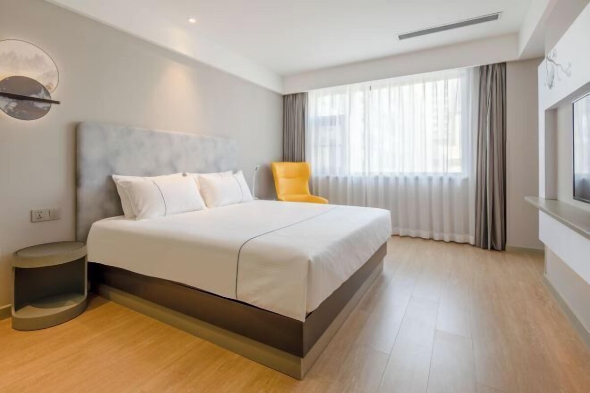 Una habitación de hotel moderna con una cama grande, una mesita de noche, una silla y un amplio ventanal con cortinas.