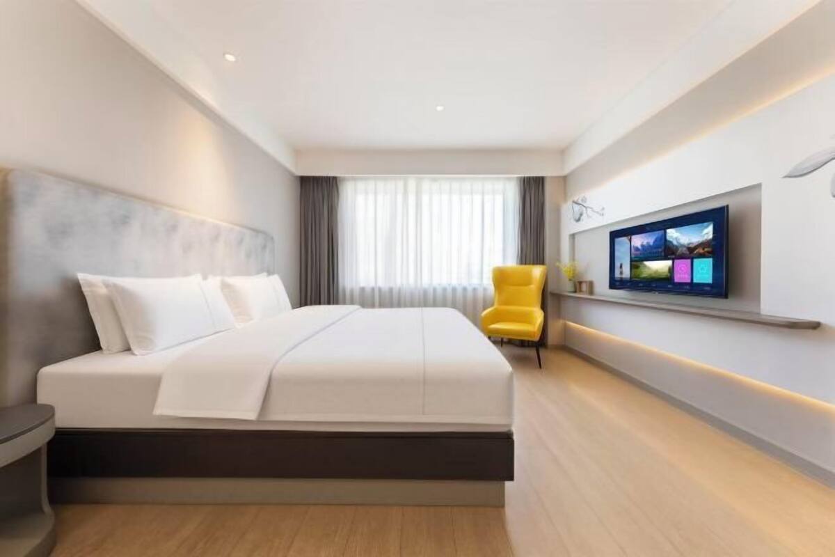 A modern hotel room with a large bed, a TV mounted on the wall, and a bright yellow chair.