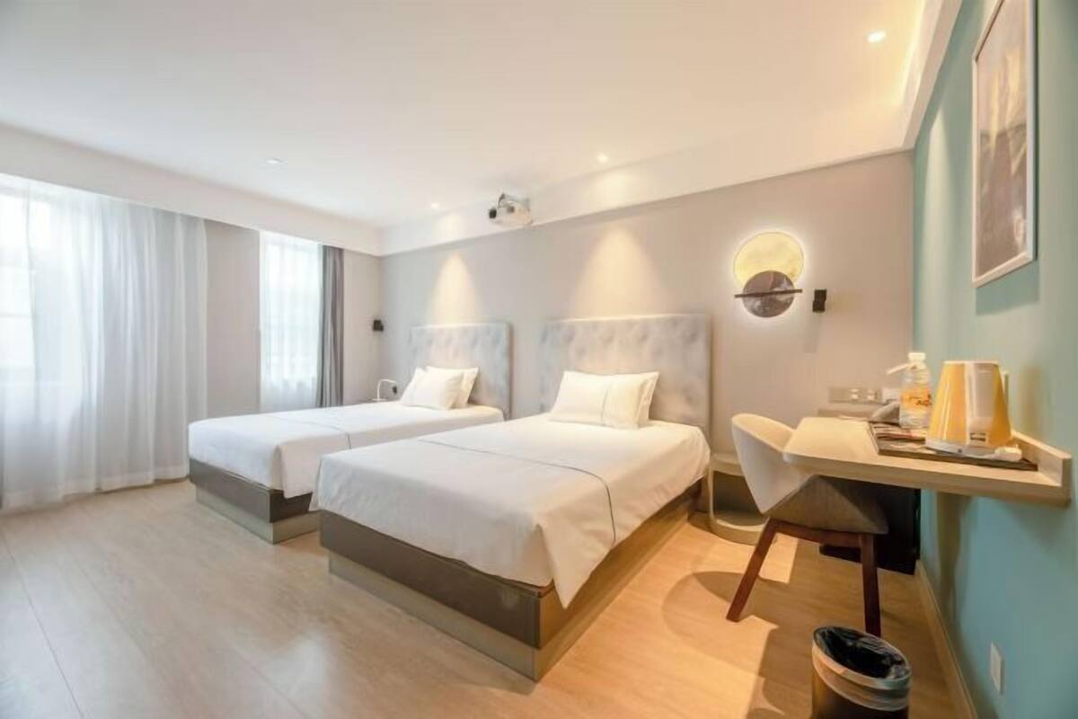 A hotel room with two beds, a desk, a chair, a trash bin, and a wall-mounted light fixture.