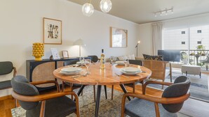 Ein Essbereich mit einem runden Holztisch, gedeckt für sechs Personen, umgeben von Korbstühlen. An den Wänden hängen zwei gerahmte Kunstwerke, ein Fernseher auf einem Schrank und durch das Fenster ist ein Balkon mit Sitzgelegenheiten im Freien zu sehen.