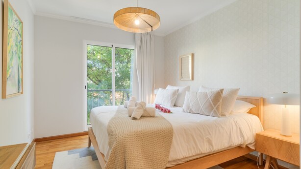 Ein Schlafzimmer mit einem großen Bett, einem Nachttisch mit Lampe, einem Fenster mit Vorhängen und Blick auf Bäume.