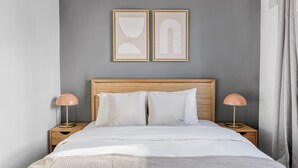 Ein Schlafzimmer mit Holzkopfstütze, zwei Nachttischlampen, einem Bett mit weißer Bettwäsche und zwei gerahmten Kunstwerken an der Wand.