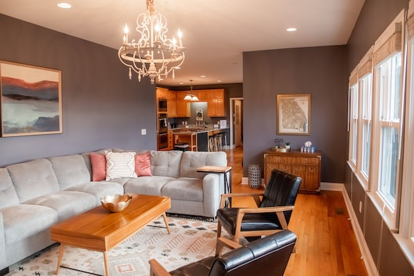 Un salon spacieux avec un grand canapé, une table basse en bois et un fauteuil en cuir. Un lustre est suspendu au plafond, et de grandes fenêtres se trouvent sur un des murs.