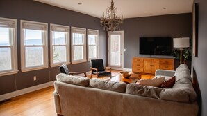 Un salón amplio con un sofá grande, una mesa de centro de madera y un televisor. Varias ventanas dejan entrar luz natural y ofrecen vistas al exterior.