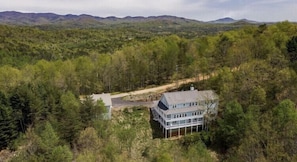 Une maison à deux étages dotée d’une grande terrasse, entourée d’une forêt dense et avec des montagnes en toile de fond.
