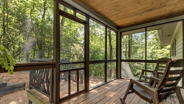 Une véranda couverte avec un plancher en bois, de grandes fenêtres et une vue sur une zone boisée.