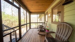 Une véranda couverte avec un plancher en bois, de grandes fenêtres et une vue sur l’extérieur.