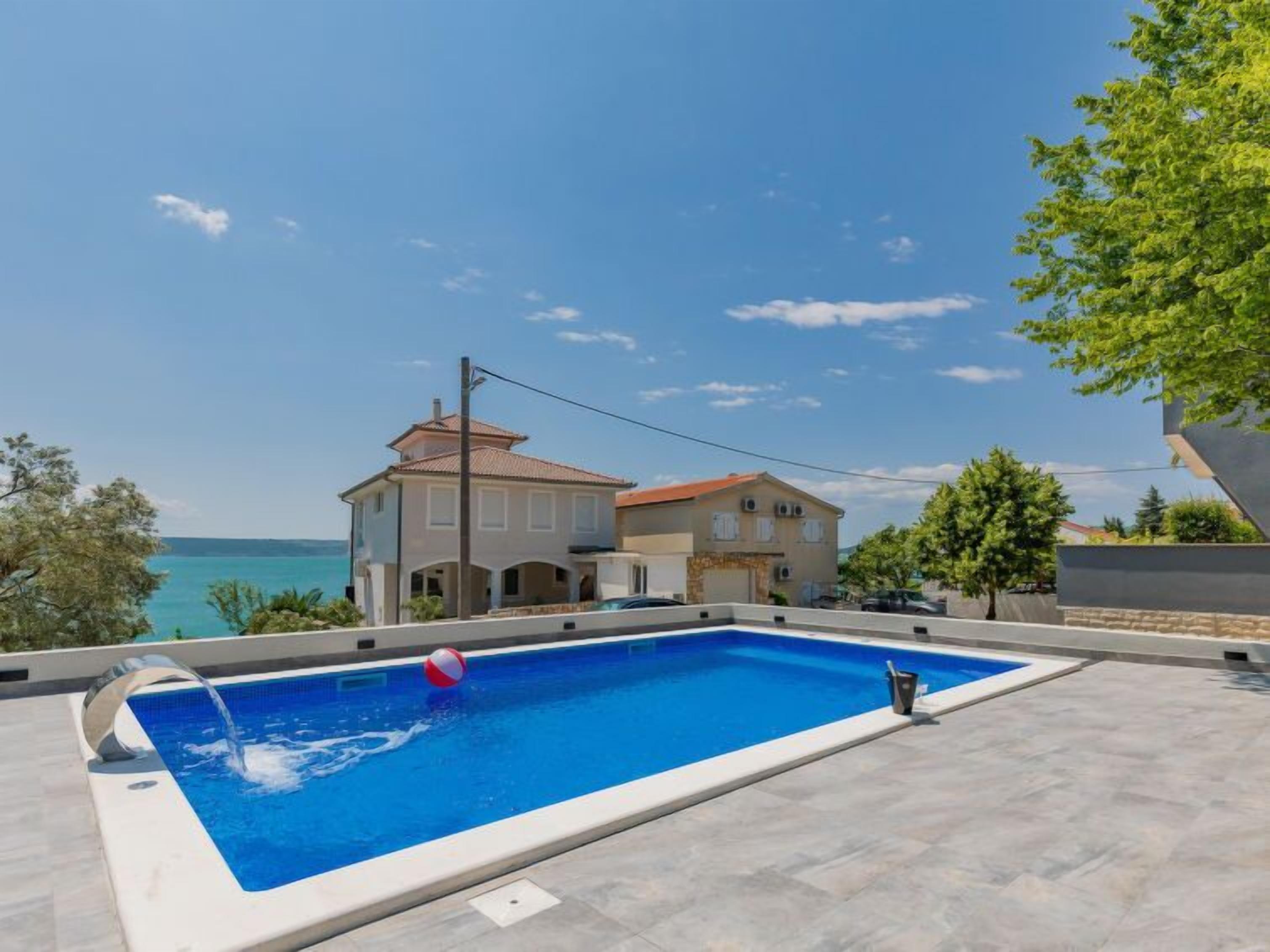 A swimming pool with a clear view of the sea, a two-story house with a red roof, and a tree to the right.