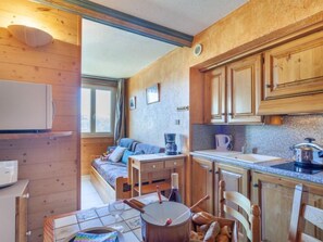 Een knus kamer in chaletstijl met een bed, een eettafel met stoelen, een keukenhoek met houten kastjes en een raam dat natuurlijk licht binnenlaat.