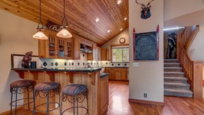 Une cuisine avec des meubles de rangement en bois, un coin bar avec des tabourets, un tableau noir avec des inscriptions et un escalier.