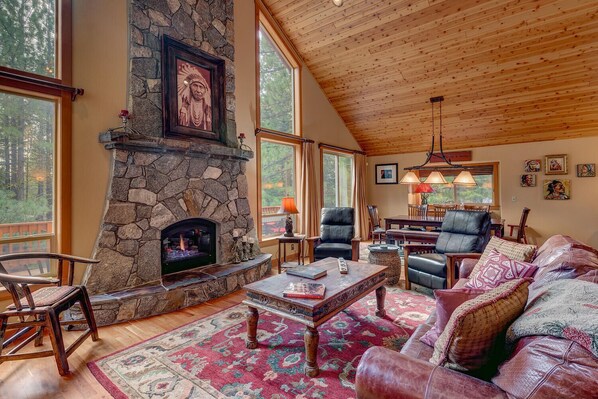 A cozy living room with a stone fireplace, leather furniture, and large windows offering a view of the outdoors. - High Country By Tahoe Getaways  - Hot Tub, EV Charger, HOA Beach and Pool (Truckee)