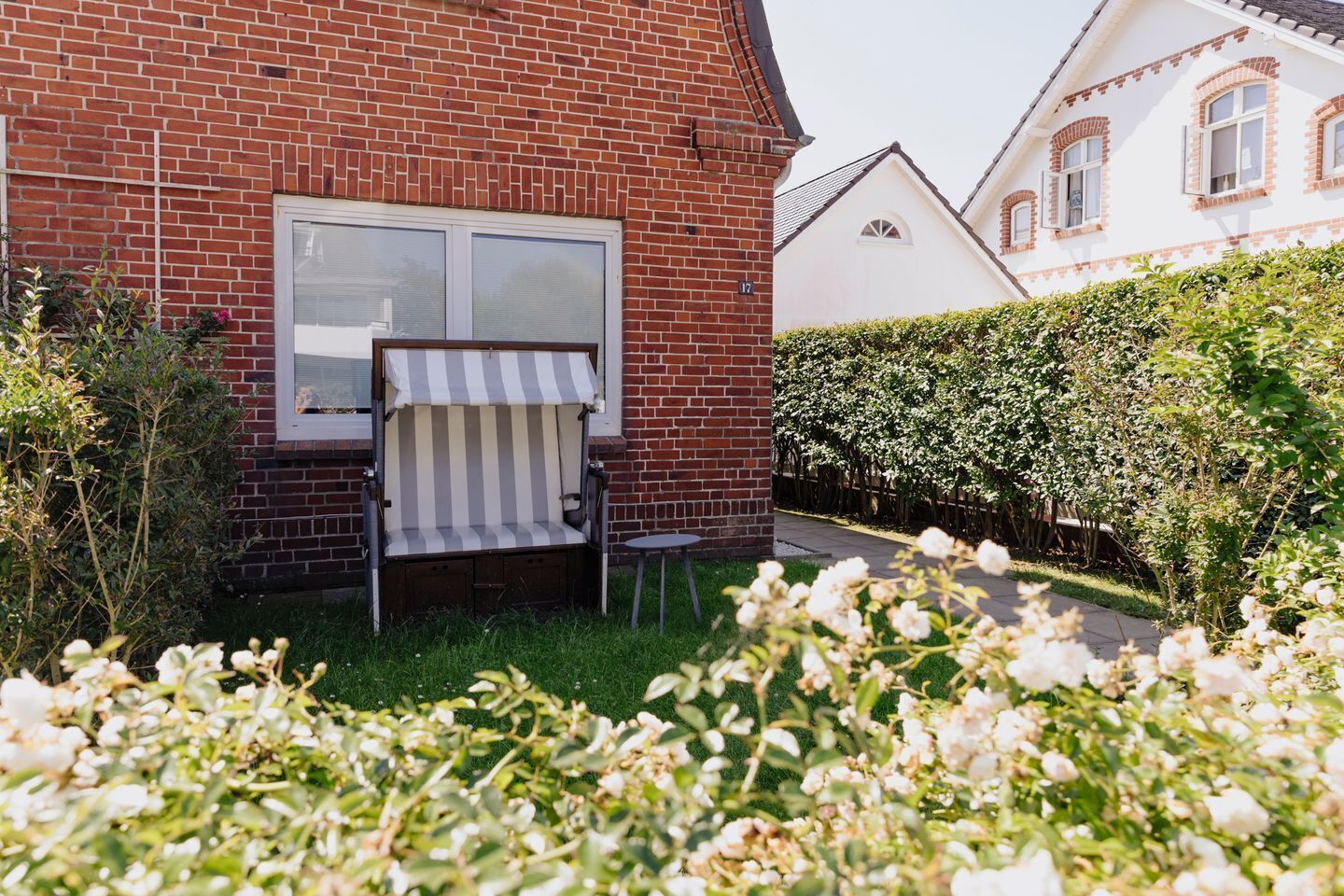 Een bakstenen huis met een gestreepte luifel boven een bank, een haag en bloeiende planten op de voorgrond.
