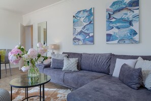 Een moderne woonkamer met een grijze bank, een glazen bijzettafeltje en een vaas met roze bloemen. Op de muur hangen twee grote schilderijen van vissen.