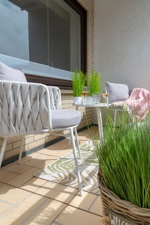 Un espace extérieur aménagé avec une chaise blanche, une petite table ornée d’une plante et d’un verre à vin, et un panier en osier contenant des plantes vertes.