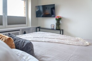 Une chambre à coucher avec un lit, un téléviseur fixé au mur, un vase avec des fleurs et une fenêtre avec des stores.