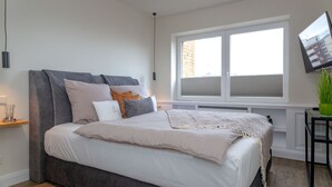 A modern bedroom with a large bed, a window with blinds, a mounted TV, and a small table with a plant. - Königsmann - Close to the beach and centrally located, modern vacation apartment with (Sylt)