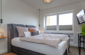 Een moderne slaapkamer met een groot bed, een raam met zonwering, een wandtelevisie en een klein tafeltje met een plant.