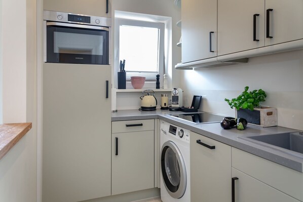Une cuisine compacte équipée d’un four encastré, d’une fenêtre, d’un lave-linge et d’un plan de travail sur lequel se trouvent divers appareils et une plante.