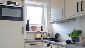 A modern kitchen with a built-in oven, microwave, and washing machine. There is a window above the sink, and a countertop with a kettle and a plant. - Königsmann - Close to the beach and centrally located, modern vacation apartment with (Sylt)