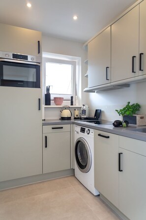 Een moderne keuken met een ingebouwde oven, magnetron en wasmachine. Boven de gootsteen is een raam en op het aanrecht staan een waterkoker en een plant.