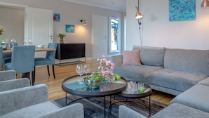 A modern living room with a grey sofa, blue chairs, a round coffee table with a flower vase, and a TV in the background.