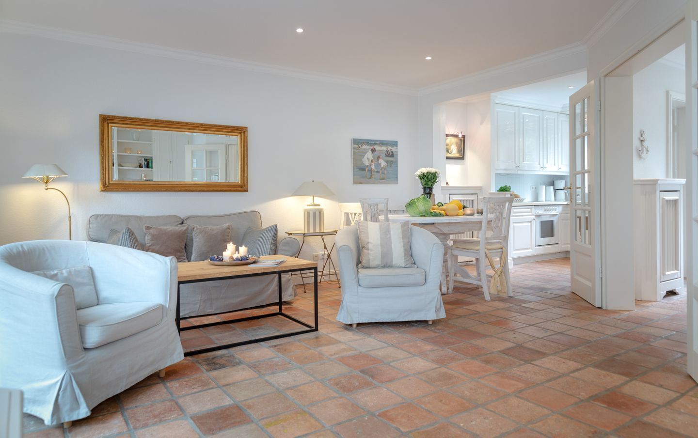 A spacious living area with a sofa, armchairs, a coffee table, and a dining table. There is a kitchen in the background with white cabinets and a tiled floor.