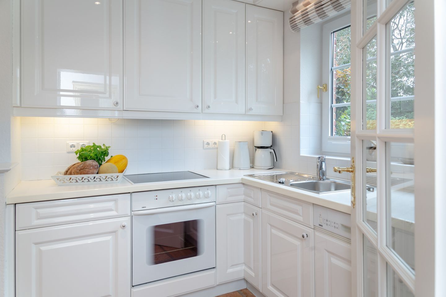 Une cuisine moderne avec des meubles blancs, un four encastré, un évier et une fenêtre donnant sur les arbres.