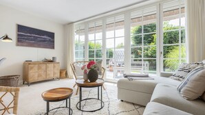 A spacious living room with a sofa, a dining table, and a chair. There is a large window with a view of greenery outside. - Tortuga - Spacious vacation home in the dreamlike captain's village of Keitum (Sylt-Ost)