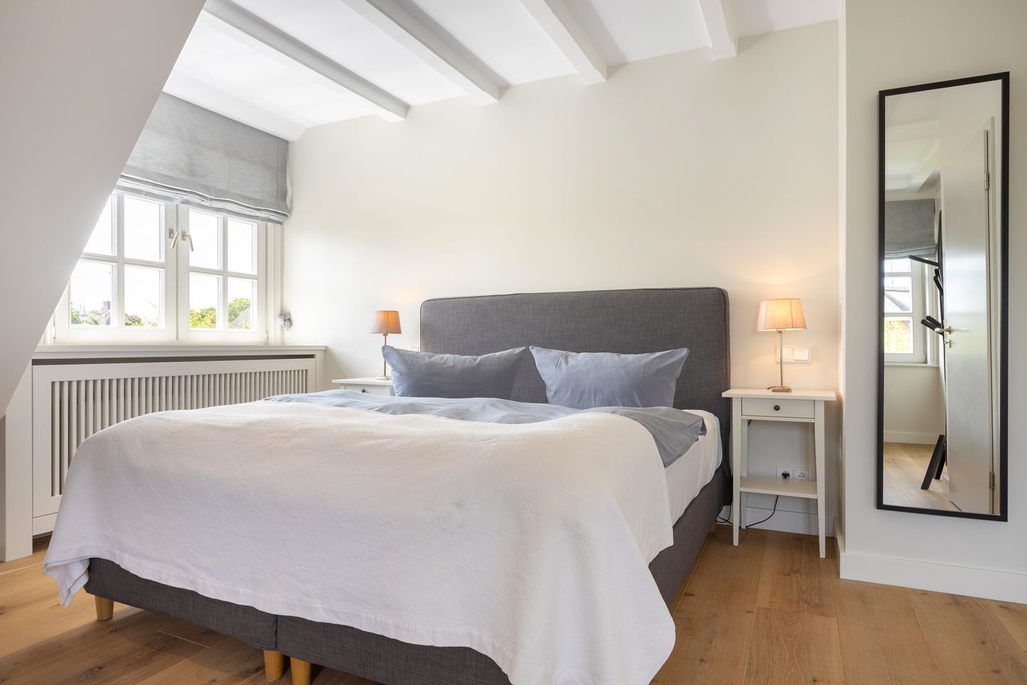 Un dormitorio con cama, dos mesitas de noche, ventana, radiador y espejo.