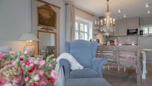 A spacious living room with a grey armchair, a white fluffy throw, and a vase of pink flowers. A large mirror and a chandelier are visible in the background.