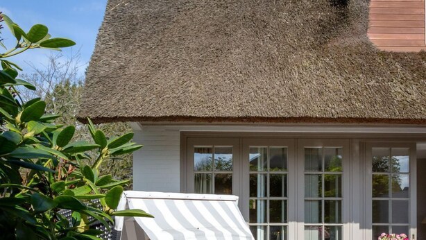 A thatched-roof house with a patio area featuring a bed, a striped canopy, and a table with chairs.
