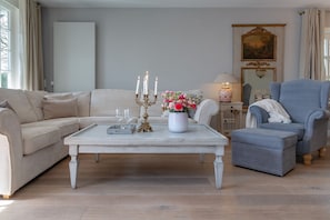 Un salon avec un canapé beige, un fauteuil gris, une table basse blanche ornée d’un vase de fleurs et de bougies, et une lampe sur une table d’appoint.