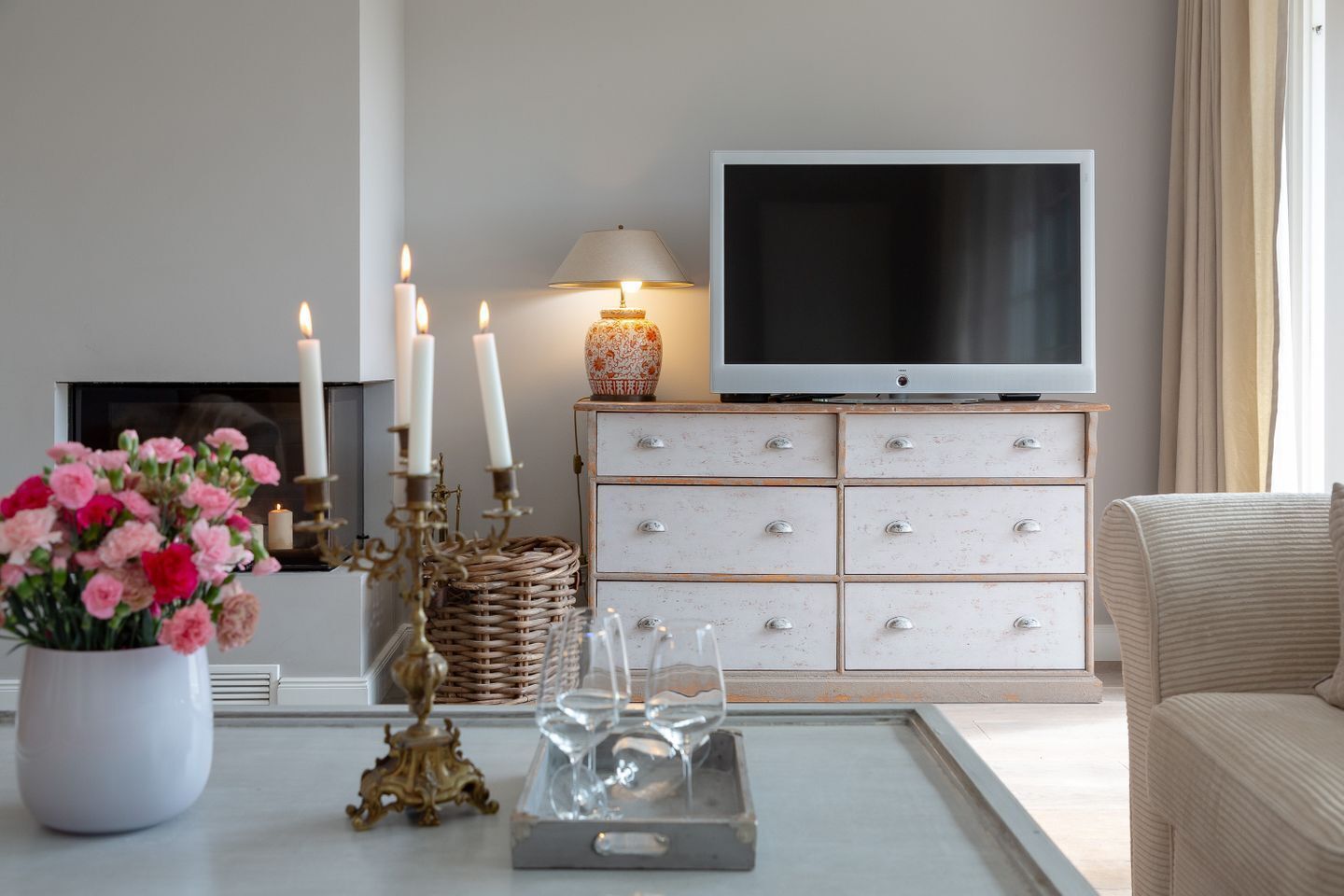 Un salon avec un meuble TV blanc, un canapé blanc, une table basse ornée d’un vase de fleurs et une cheminée.