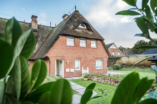 Une maison en briques avec un toit de chaume, une porte blanche et un petit jardin avec un chemin menant à l’entrée.