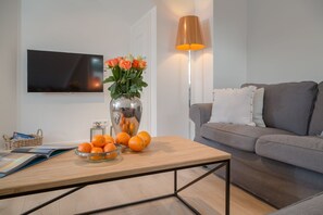Un salon moderne avec un canapé gris, une table basse en bois ornée d’oranges et d’un vase de fleurs, et un lampadaire.