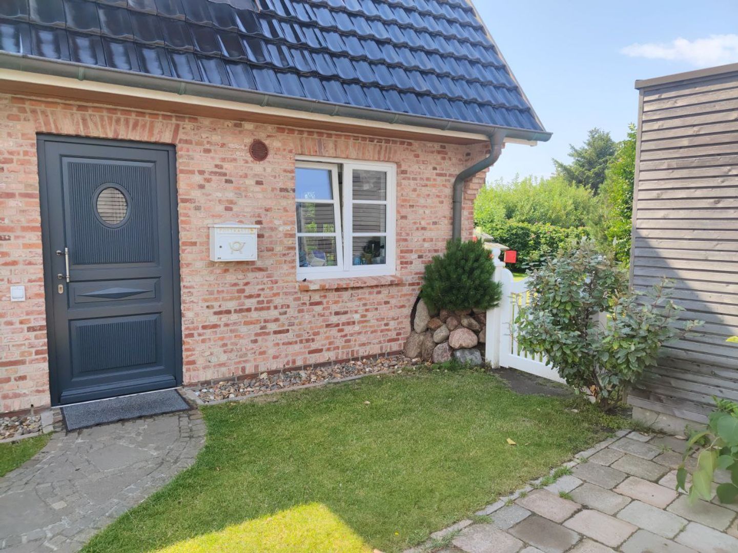 Une maison en briques avec une porte bleu foncé, une petite fenêtre et un mur de pierres surmonté d’une clôture blanche.
