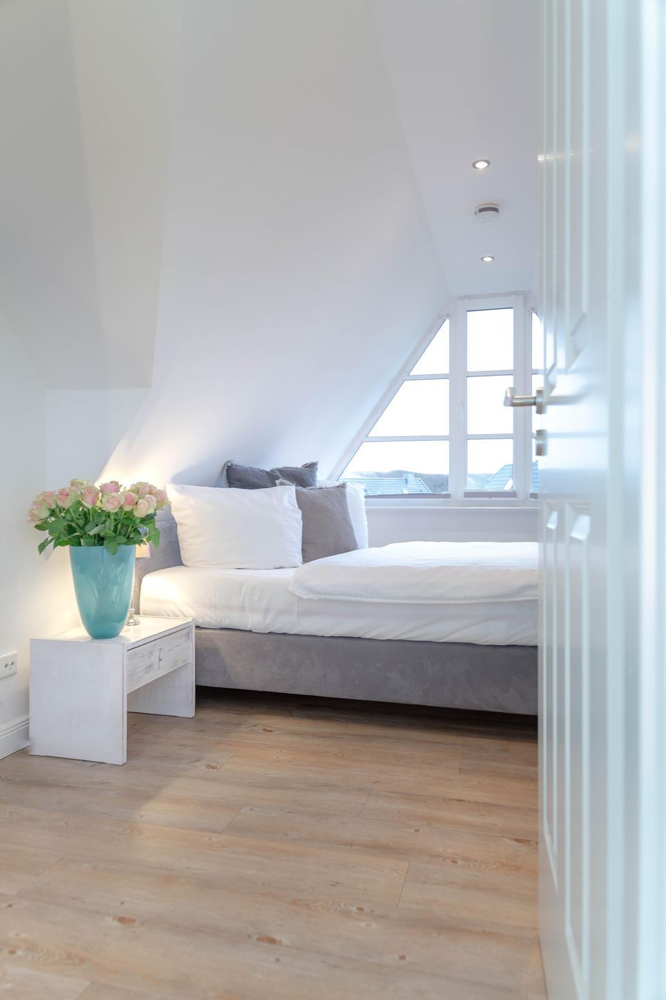 A bedroom with a sloped ceiling, a bed with white bedding, a window with blinds, and a vase with flowers on a bedside table.