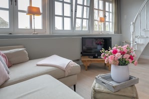 Una sala de estar con un sofá, un televisor sobre un mueble de madera y un jarrón de flores sobre una mesa.