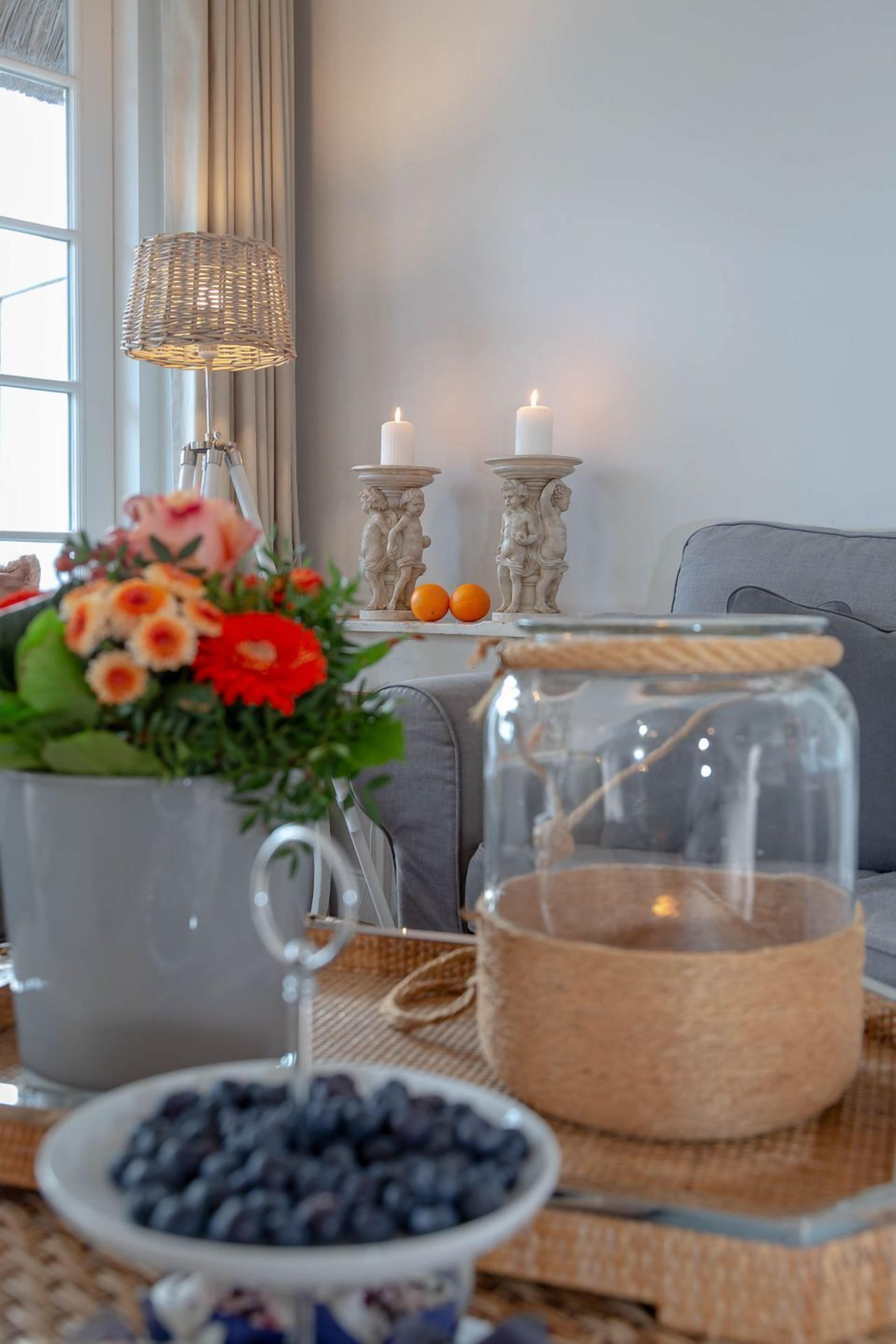 Ein gemütliches Wohnzimmer mit einer Lampe, Kerzen, einer Blumenvase, einem Glasbehälter und einer Schale Blaubeeren auf einem gewebten Tisch.