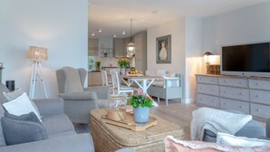 Un salon moderne avec un canapé gris, un coin repas avec une table et des chaises blanches, une cuisine en arrière-plan et un téléviseur sur le côté droit.