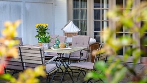 Une terrasse extérieure avec une table et des chaises en bois, un fauteuil en osier, une petite table avec une lampe et un pot de fleurs contenant des fleurs jaunes.